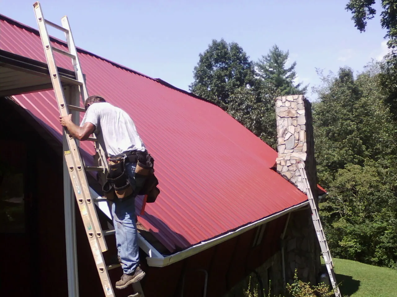 Roofer on a ladder beside a red metal roof and stone chimney — Soffit & Fascia Repair by Roofing Brothers in Providence Forge